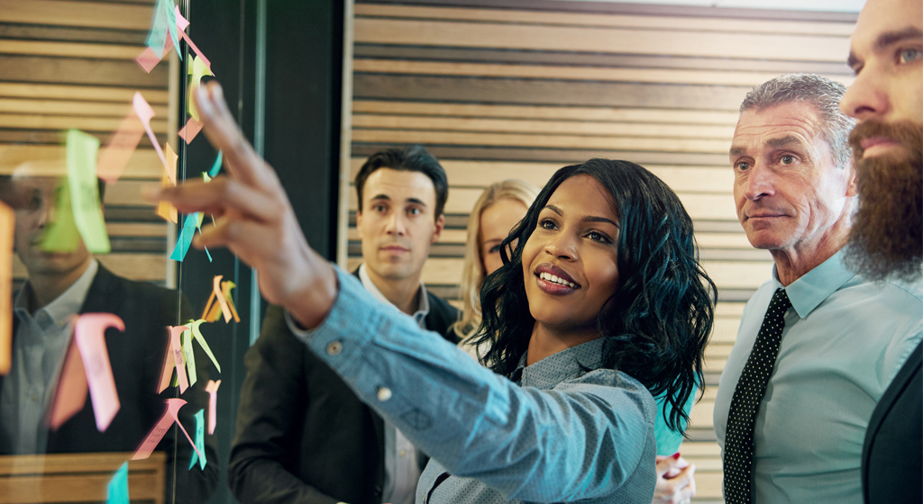 woman pointing to notes on a wall with people gathered around in a group looking at the notes.