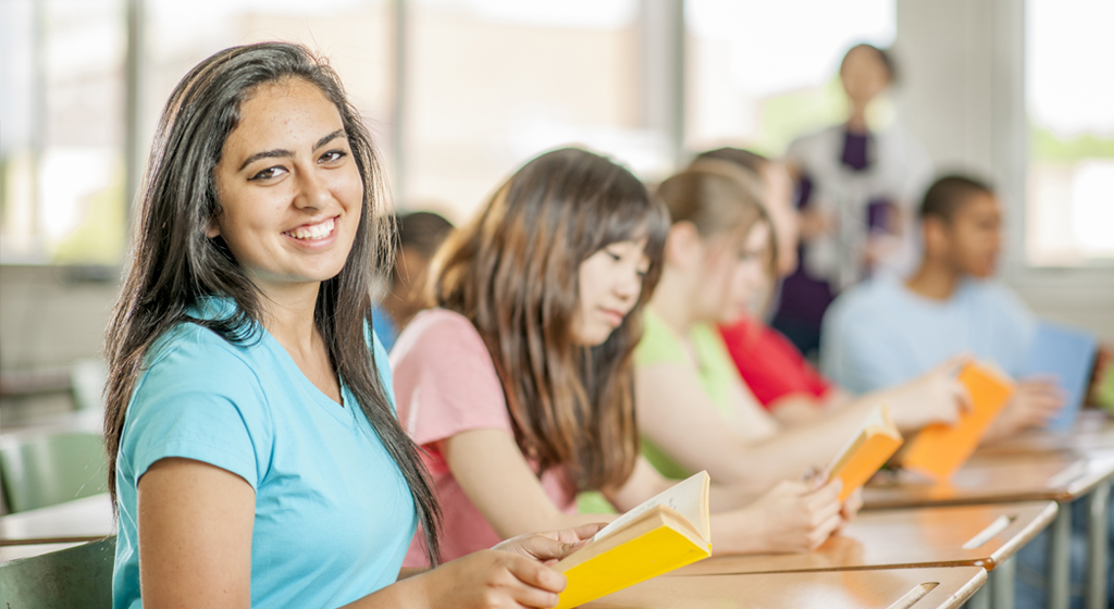 woman sitting at a desk in a classroom faces the camera and smiles