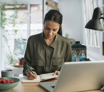 woman writing in a notebook at table in front of a laptop computer