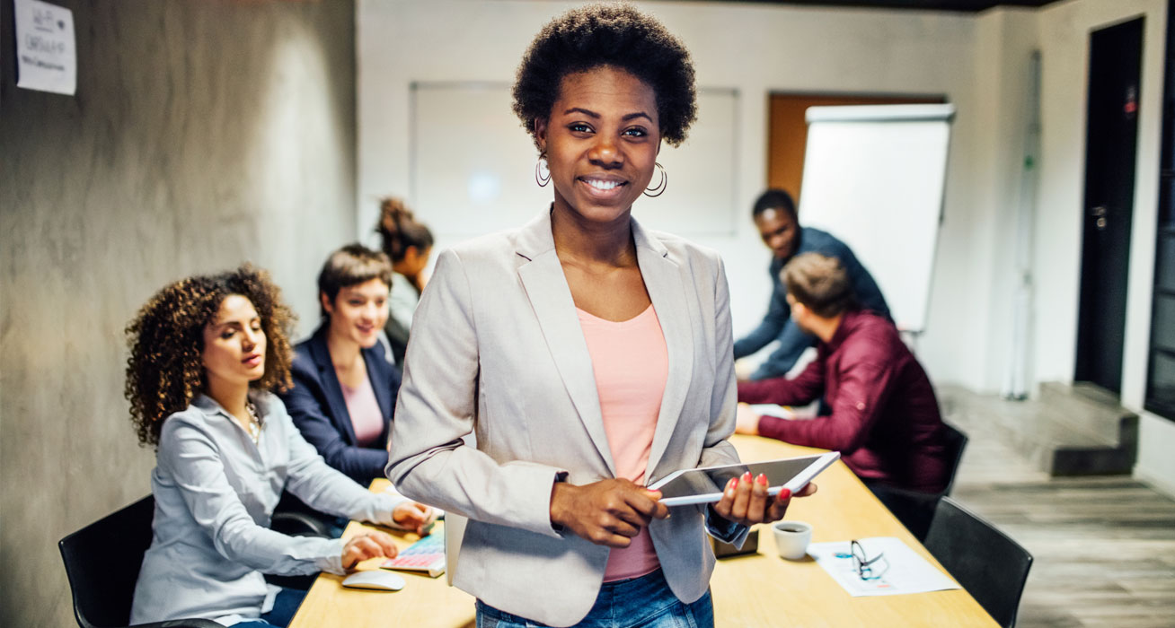 people at a conference table, women stands and faces camera holding a tablet