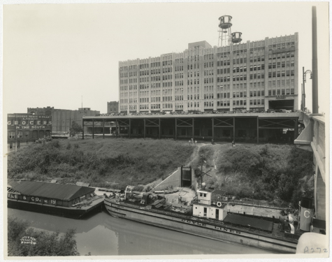 OMB (M & M Building at that time) shown when it was an industrial center. Photo shows a boat and barge docking to unload goods.