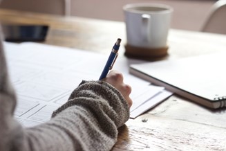 Someone holding pencil writing down notes with a coffee mug on table.